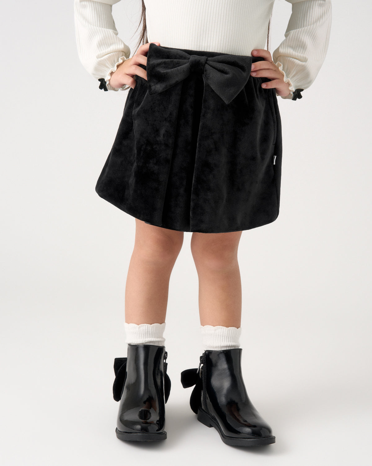 A child wearing a black velvet skirt with a large bow at the waist, paired with a white ribbed sweater, white socks, and black glossy ankle boots, shown from the waist down against a light gray studio background.