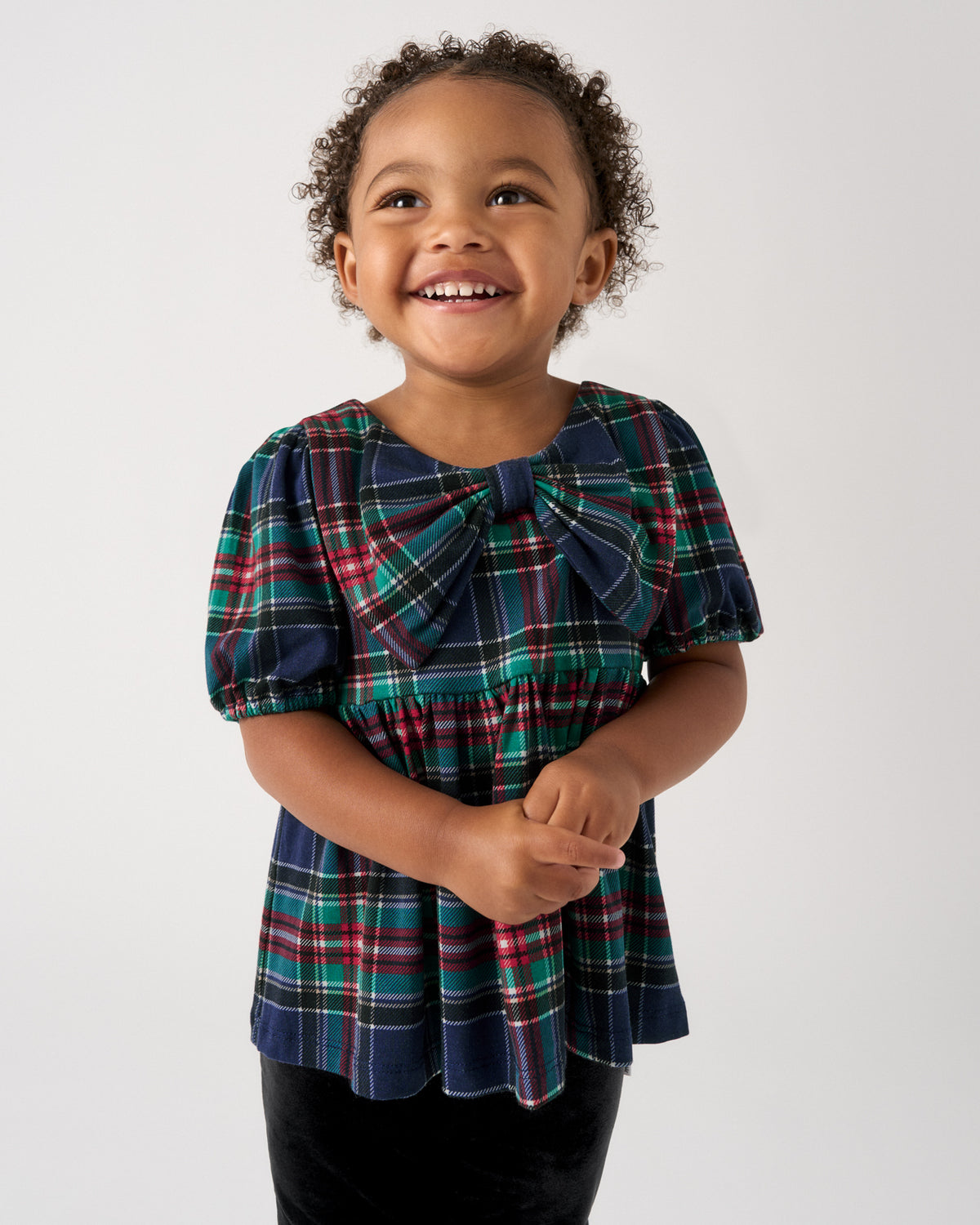 A young child with curly hair smiling, wearing a navy blue plaid top with a large bow at the neckline and puff sleeves, paired with black pants, against a plain light gray background.