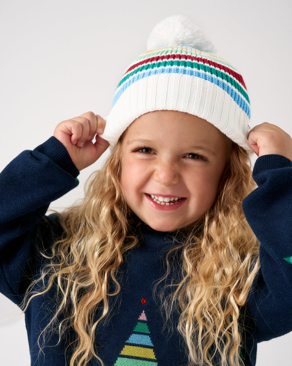 A young child with long blonde hair wearing a white knit pom-pom beanie with colorful horizontal stripes and a navy sweater, posing against a light gray background.