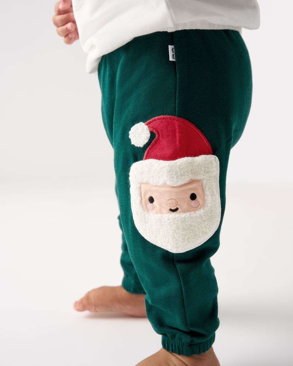 A close-up of a child wearing dark green fleece pants with a large applique of a Santa Claus face on the leg, paired with a white top against a light gray studio background.