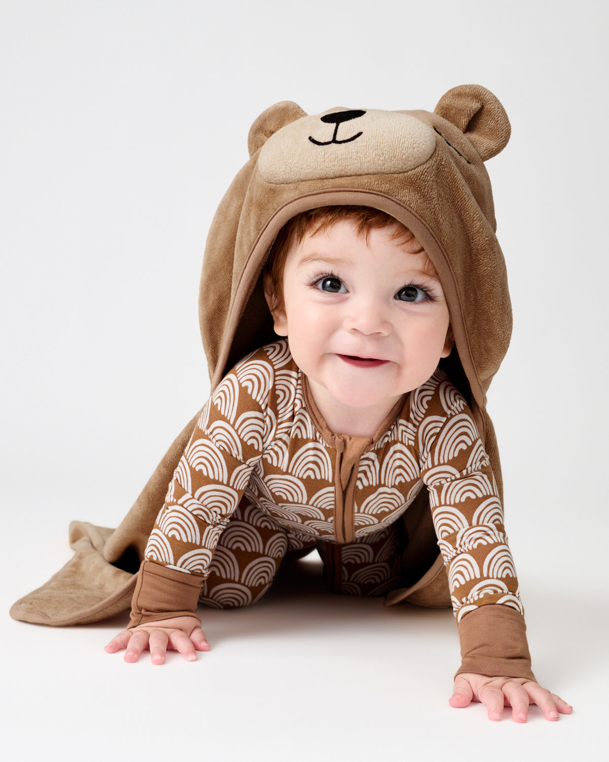 A baby crawling on a light gray studio floor wearing a brown hooded one-piece outfit with a bear face on the hood and a beige and brown wave pattern on the body.