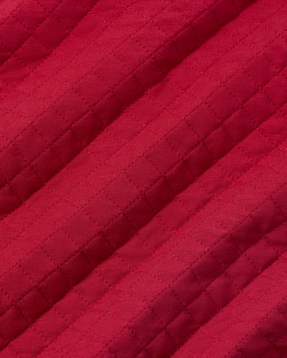 A close-up of a quilted fabric in a rich magenta-red color, showing the diamond stitching pattern and diagonal folds.