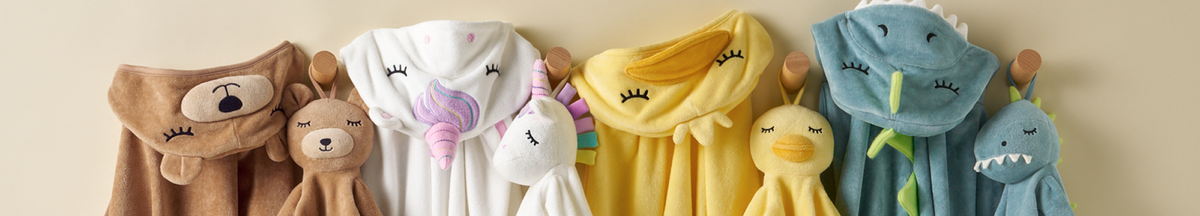 A row of plush animal and monster hooded towels hanging on wooden pegs against a beige wall. The towels include a brown bear, a white unicorn with pink and purple accents, a yellow duck, and blue shark and monster designs.