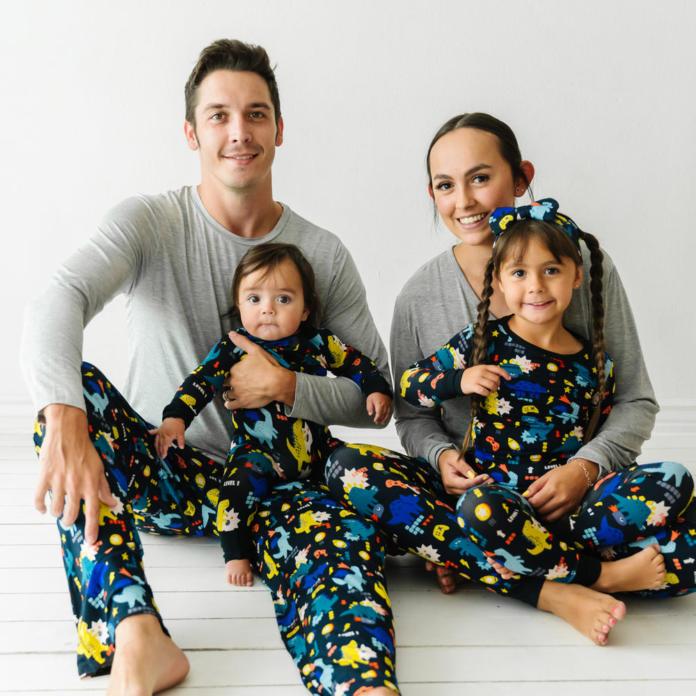 Family of four posing together wearing coordinating Next Level Dinos pajamas. Dad is wearing Next Level Dinos men's pajama pants paired with a Heather Gray men's pajama top. mom is wearing Next Level Dinos women's pajama pants paired with a Heather Gray women's pajama top. Their children are wearing matching Next Level Dinos pajamas in two piece and zippy styles paired with a luxe bow headband