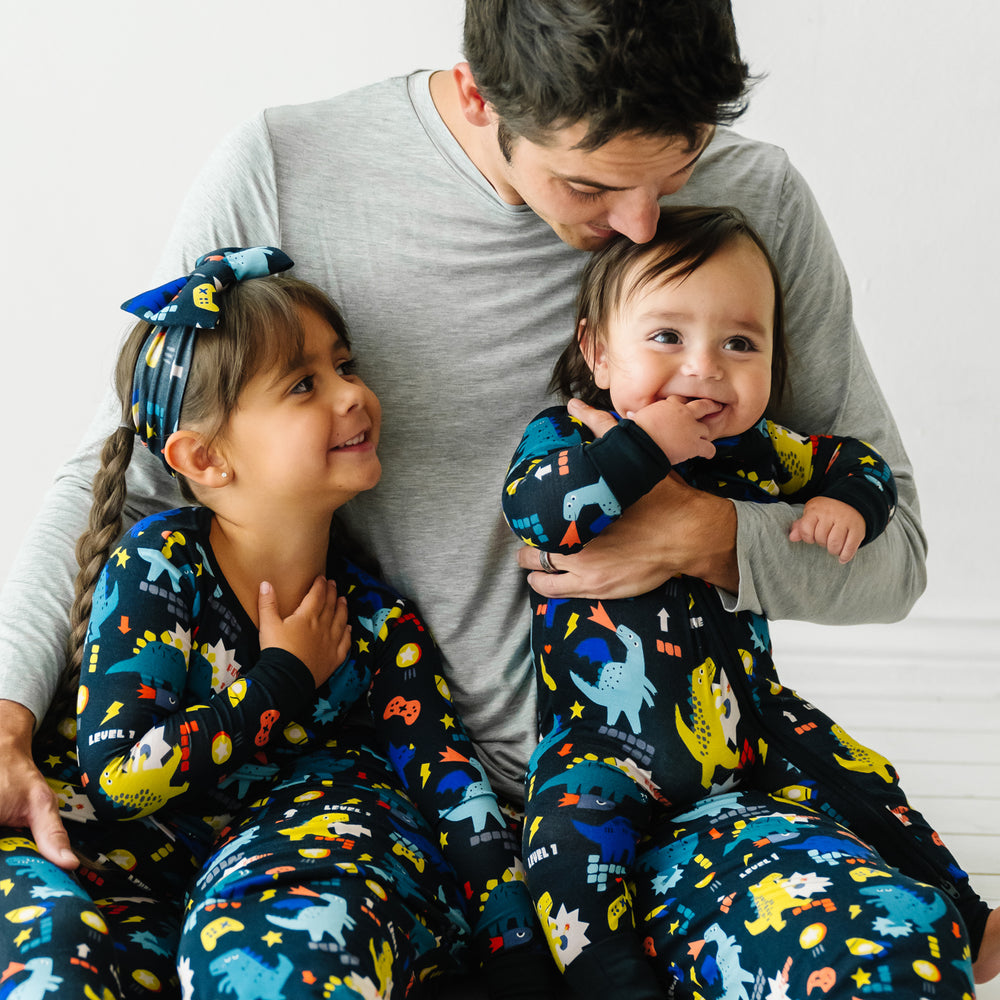 Dad and his two children sitting together. Dad is wearing Next Level Dinos men's pajama pants paired with a men's Heather Gray pajama top. Children are wearing Next Level Dinos pajamas in two piece and zippy styles paired with a matching luxe bow headband