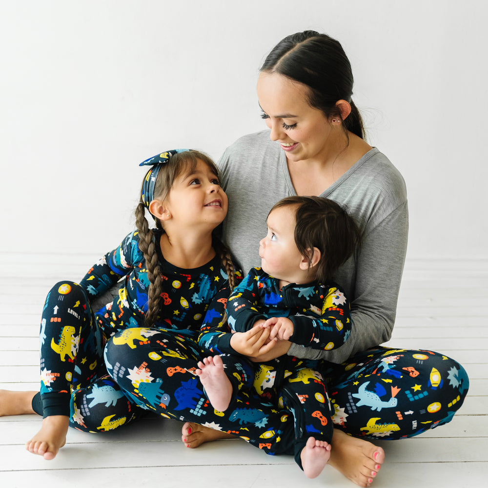 Mom and her two children sitting together. Mom is wearing Next Level Dinos women's pajama pants paired with a women's Heather Gray pajama top. Children are wearing Next Level Dinos pajamas in two piece and zippy styles paired with a matching luxe bow headband