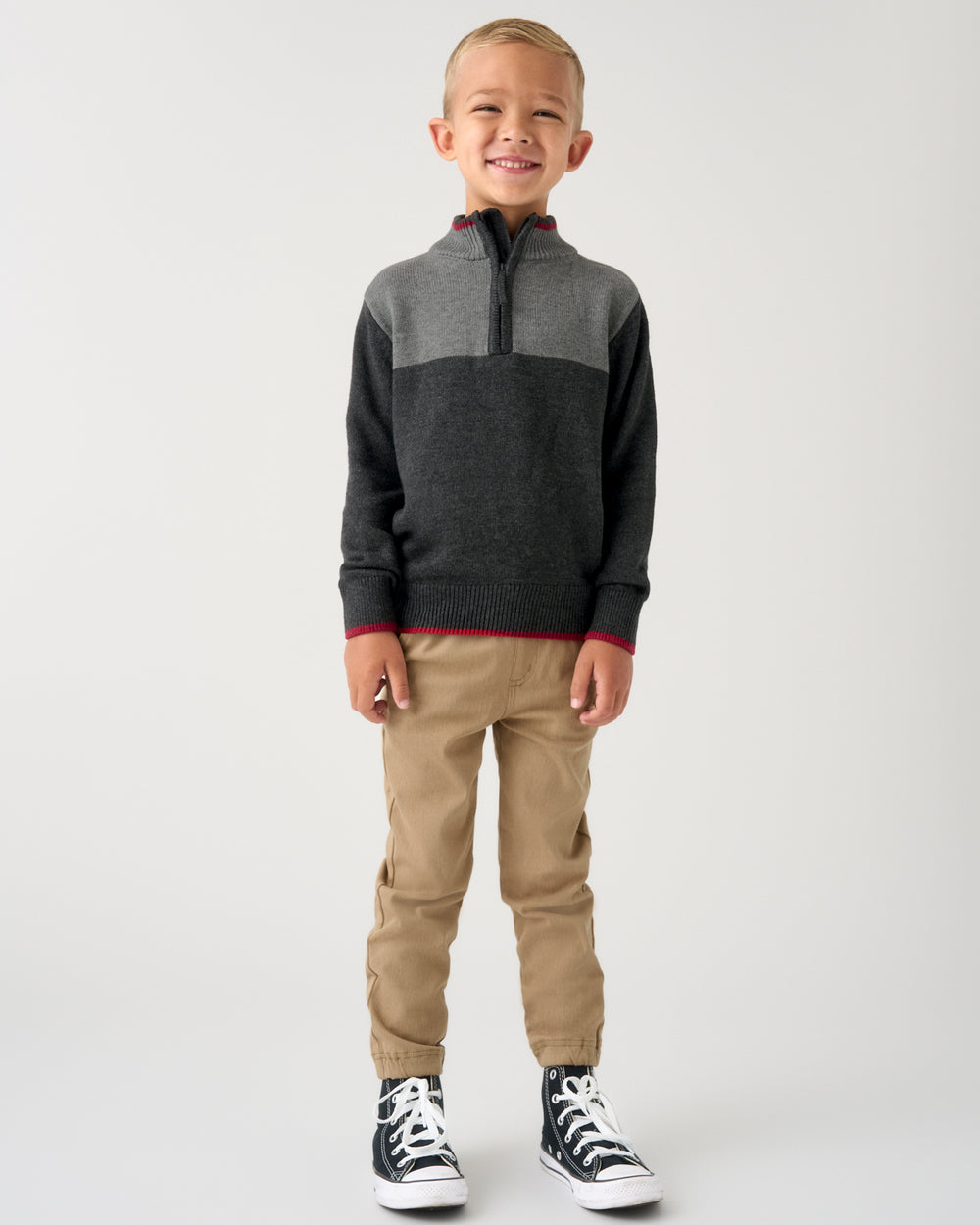 A young boy stands against a light gray studio background wearing a color-blocked quarter-zip sweater, tan pants, and black-and-white sneakers.