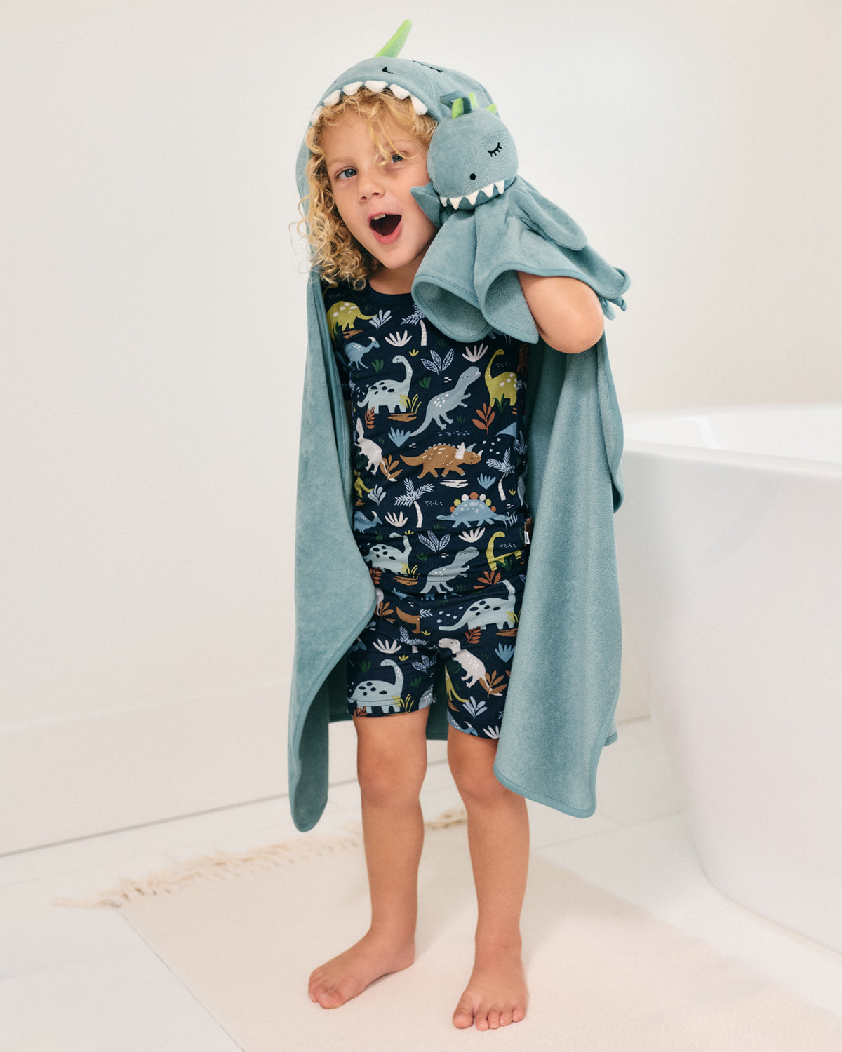 A young child wearing a navy blue dinosaur-print pajama set and wrapped in a light blue dinosaur hooded towel, standing barefoot in a bright bathroom.