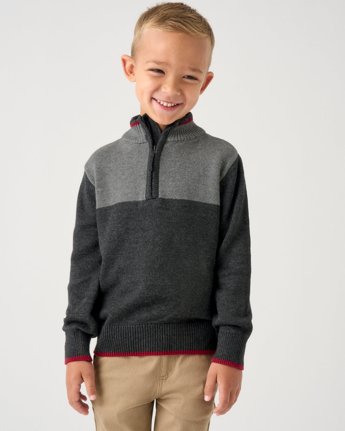 A young boy smiling while wearing a gray and dark charcoal knit quarter-zip sweater with red trim, paired with khaki pants, against a light gray background.