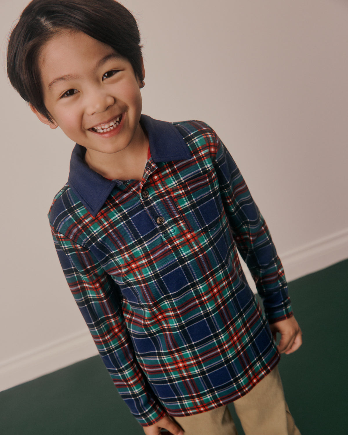 A smiling young boy wearing a long-sleeve plaid polo shirt with a navy blue collar, featuring red, green, and white stripes on a navy base, paired with khaki pants.
