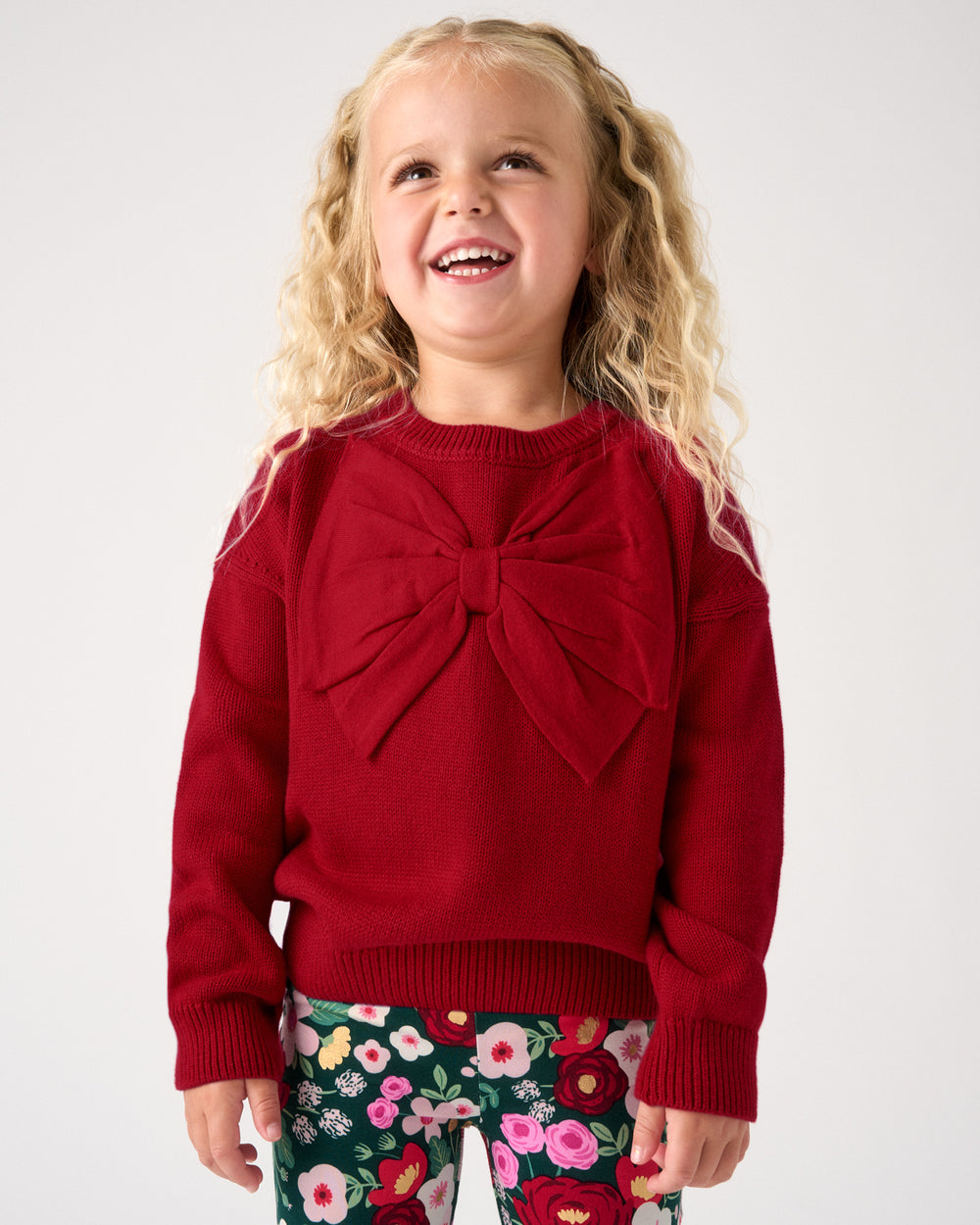 A young girl with blonde curly hair smiling, wearing a red knit sweater with a large bow on the chest and green floral leggings, against a plain light gray background.