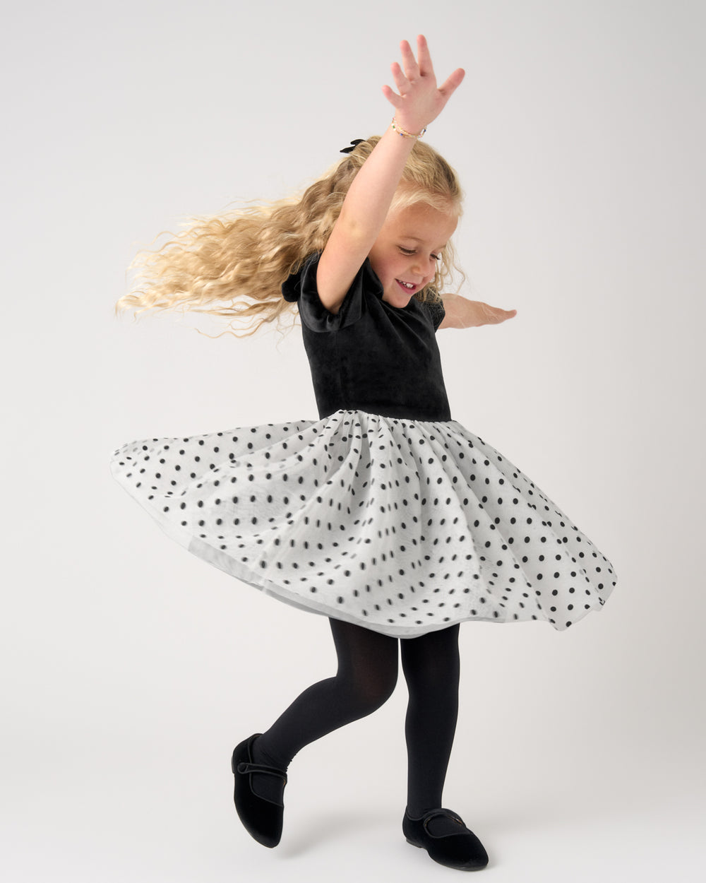 A young girl with long blonde hair is captured mid-spin, wearing a black short-sleeve top and a white flared skirt with black polka dots, paired with black tights and black shoes, against a plain light gray studio background.