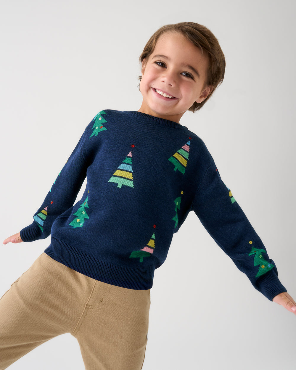 A smiling young boy wearing a navy blue crewneck sweater with colorful Christmas tree patterns and khaki pants, posing against a light gray studio background.