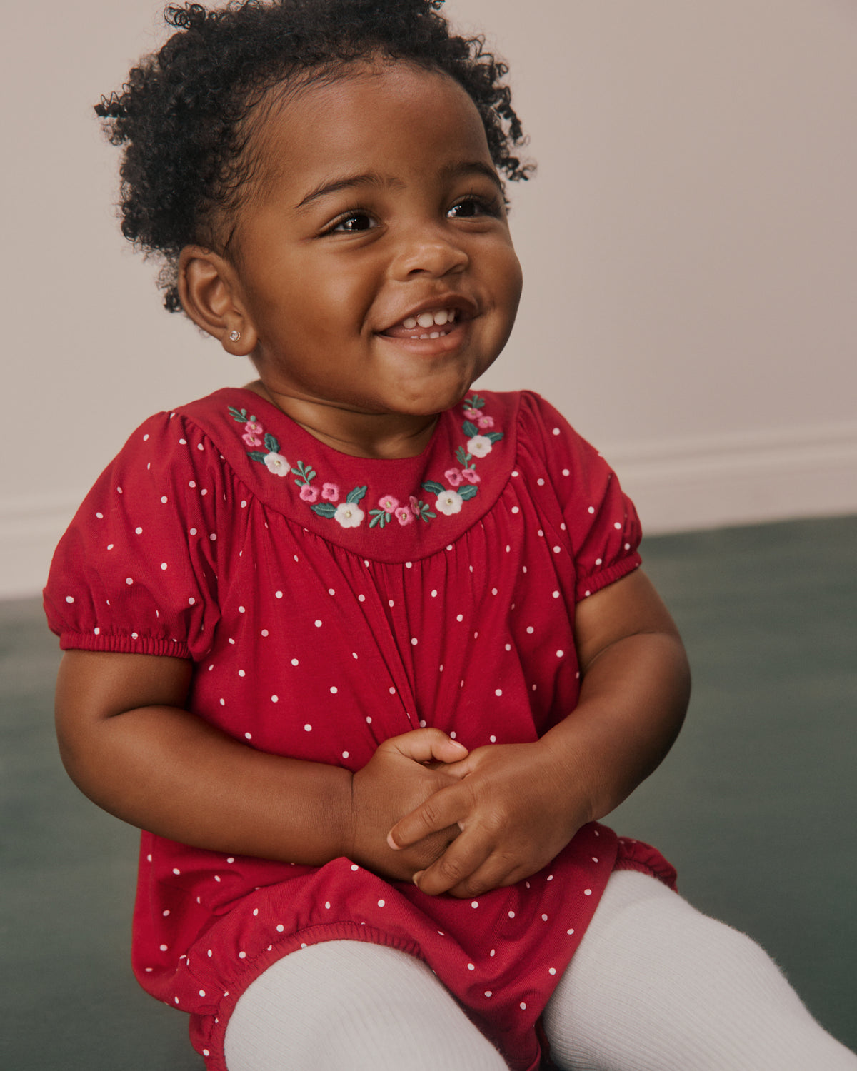 A smiling toddler with curly dark hair wearing a red short-sleeve dress with white polka dots and a floral embroidered neckline, paired with white tights, sitting indoors against a neutral wall and dark green floor.