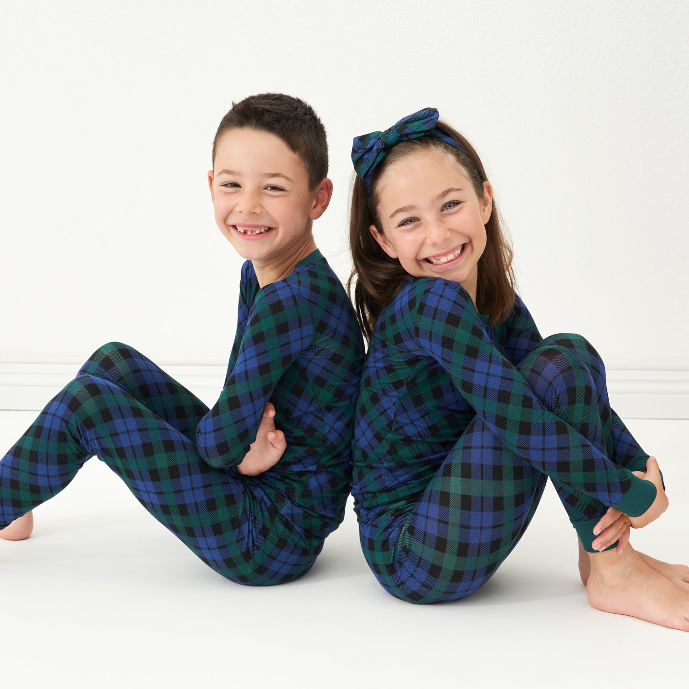 Two children sitting back to back wearing matching Emerald Plaid two-piece pajama sets
