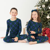 Two children sitting in front of a tree and presents wearing matching Emerald Plaid two-piece pajama sets