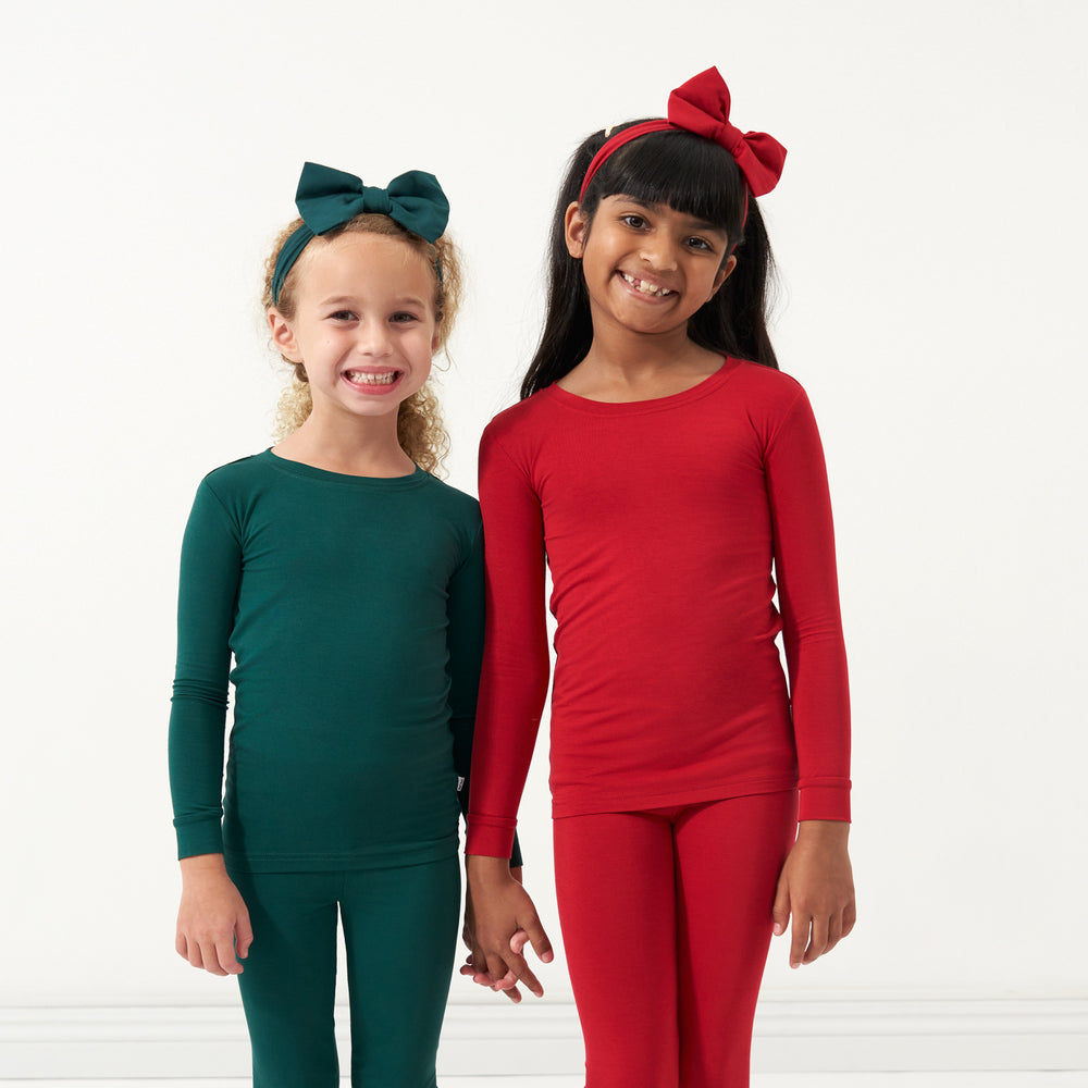 Two children wearing coordinating Emerald and Holiday Red two-piece pajama sets and luxe bow headbands