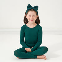 Child sitting on the ground wearing an Emerald two-piece pajama set and matching luxe bow headband