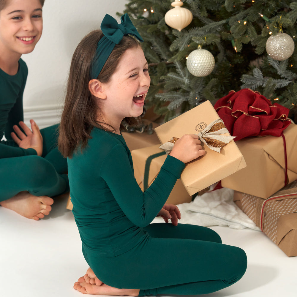 Child sitting in front of a tree wearing an Emerald two-piece pajama set and matching luxe bow headband