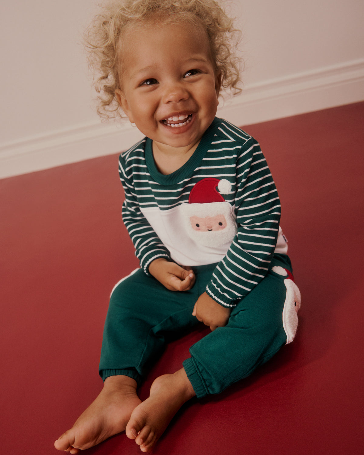 A smiling toddler with curly hair sits barefoot on a red floor against a light wall, wearing a dark green and white striped long-sleeve top with a Santa Claus face graphic and dark green pants.