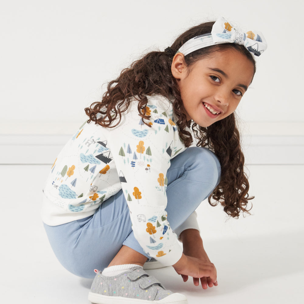 Child kneeling down wearing a Let's Explore printed crewneck sweatshirt and luxe bow headband with coordinating leggings