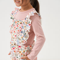 Close up image of a child wearing Mauve Meadow printed ruffle overalls over a Mauve classic tee