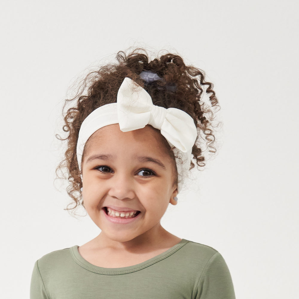 Click to see full screen - Child wearing an Ivory luxe bow headband paired with a Moss classic tee