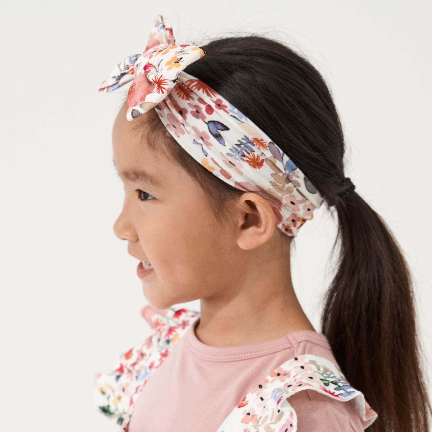 Alternate profile view of a child wearing a Mauve Meadow printed luxe bow headband. She is pairing her headband with matching Mauve Meadow printed ruffle overalls with a Mauve play tee