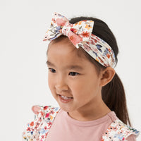 Profile view of a child wearing a Mauve Meadow printed luxe bow headband. She is pairing her headband with matching Mauve Meadow printed ruffle overalls with a Mauve play tee