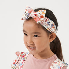 Profile view of a child wearing a Mauve Meadow printed luxe bow headband. She is pairing her headband with matching Mauve Meadow printed ruffle overalls with a Mauve play tee
