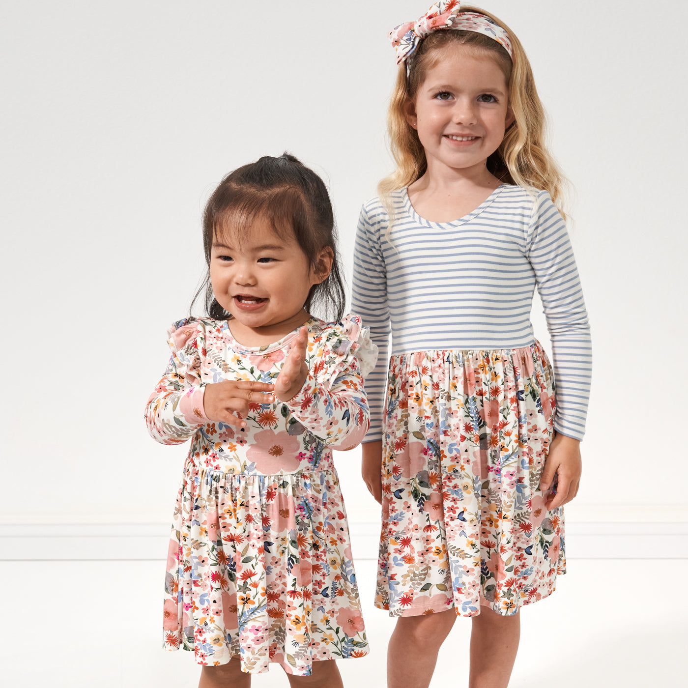 Two children posing together wearing Little Sleepies play styles. One child is wearing a Mauve Meadow printed twirl dress with bodysuit, the other child is wearing a Mauve Meadow and Ivory and Fog Striped twirl dress paired with a matching Mauve Meadow luxe bow headband