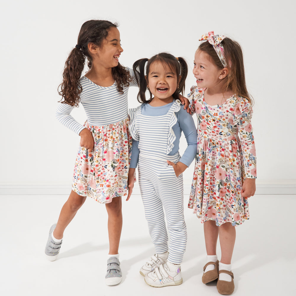 Three children posing together wearing various play styles. One child is wearing a Mauve Meadow and Ivory Fog striped twirl dress. Another child is wearing a Fog bodysuit paired with Ivory and Fog flutter overalls. The last child is wearing a Mauve Meadow printed twirl dress paired with a matching Mauve Meadow printed luxe bow headband