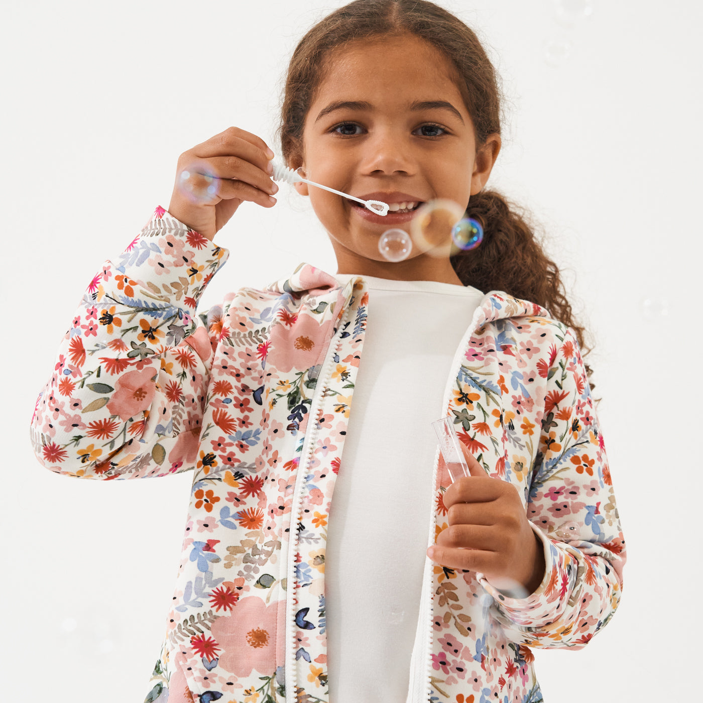 Child blowing bubbles wearing a Mauve Meadow printed peplum hoodie over an Ivory classic tee