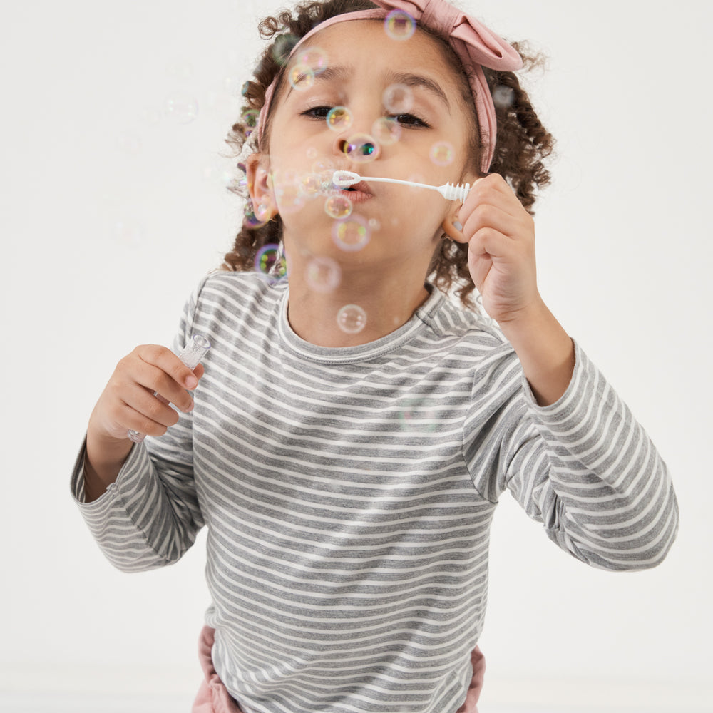Child blowing bubbles wearing a Heather Gray and Ivory Stripe classic tee paired with Mauve Blush paperbag joggers paired with a Mauve Blush luxe bow headband