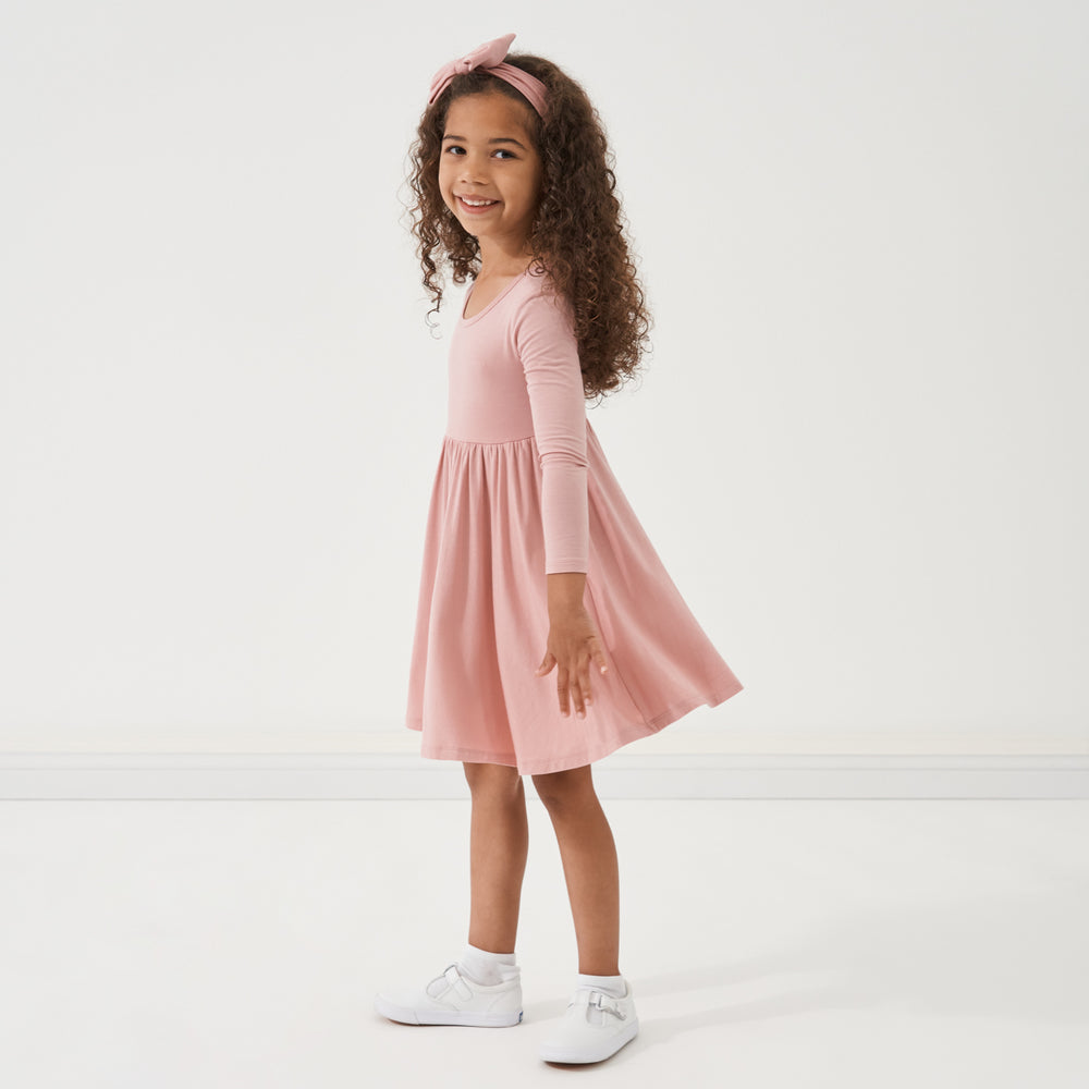 Click to see full screen - Profile view of a child wearing a Mauve Blush twirl dress paired with a matching luxe bow headband