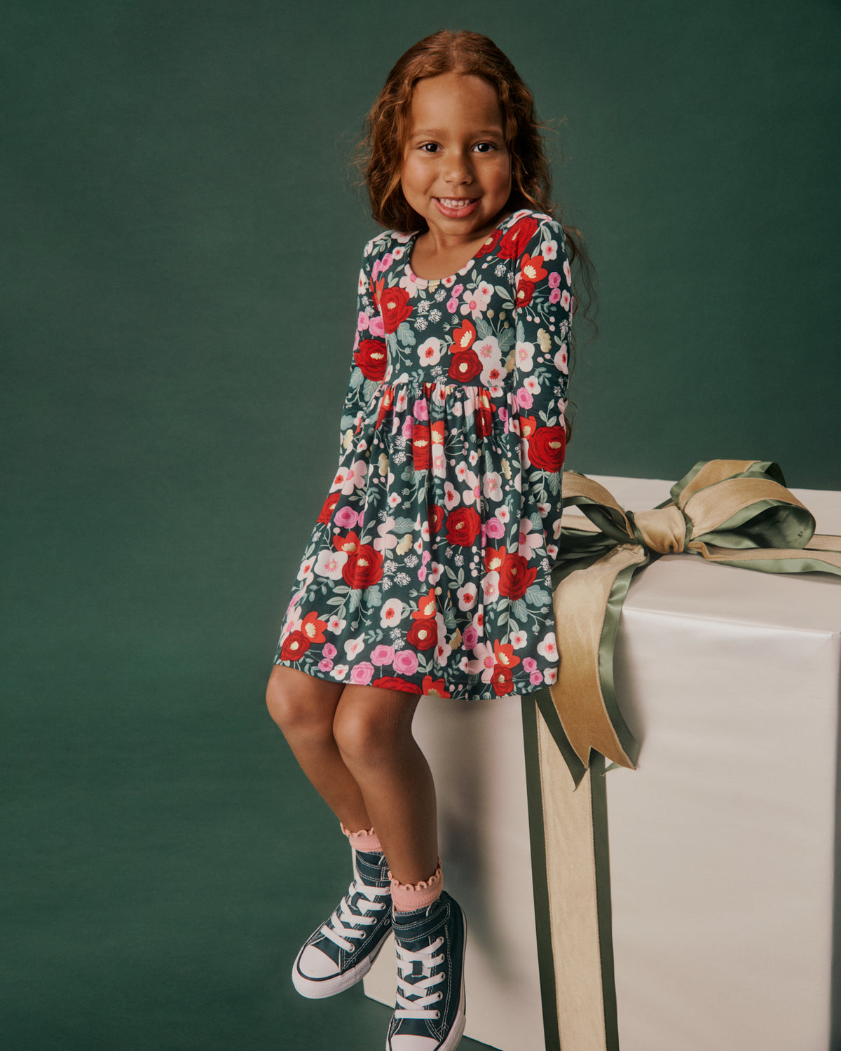 A young girl with long brown hair sits on a large white gift box with a gold ribbon, wearing a dark green long-sleeve dress with a colorful red, pink, and white floral pattern, paired with navy high-top sneakers.
