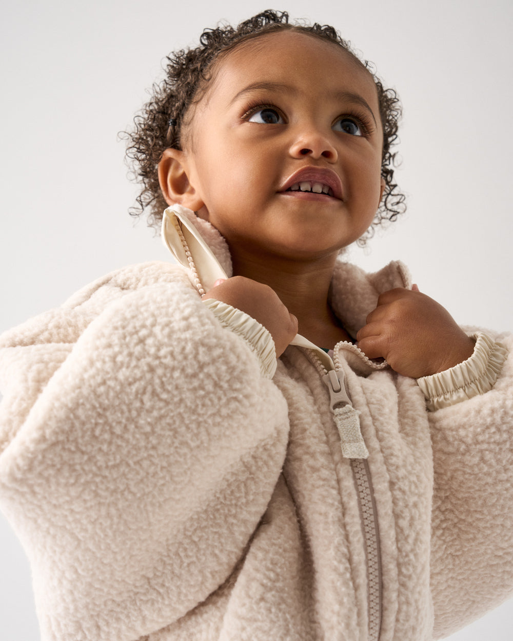 A close-up of a toddler wearing a cream-colored sherpa fleece zip-up jacket, looking upward against a plain light gray background.