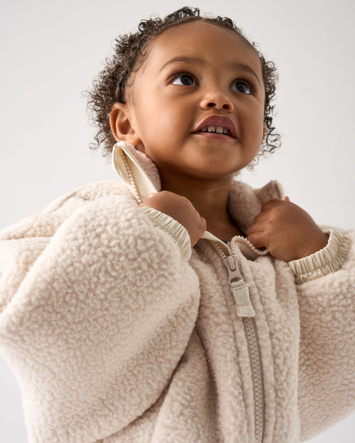 A close-up of a toddler wearing a cream-colored sherpa fleece zip-up jacket, looking upward against a plain light gray background.