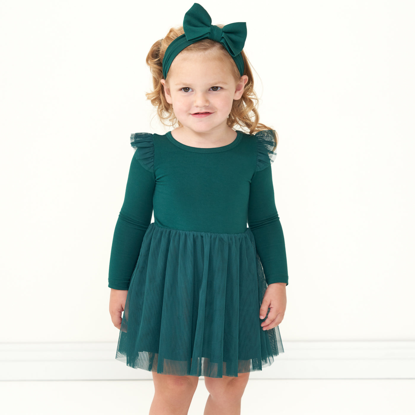 Child posing wearing an Emerald flutter tutu dress with bloomer paired with an Emerald luxe bow headband