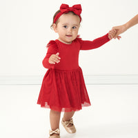 Child holding their mother's hand wearing a Holiday Red flutter tutu dress with bloomer paired with a matching Holiday Red luxe bow headband