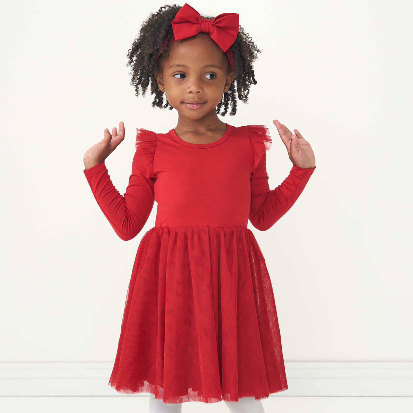 Child posing wearing a Holiday Red flutter tutu dress paired with a matching Holiday Red luxe bow headband