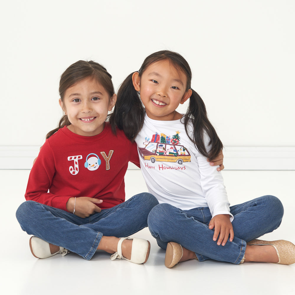 Two children sitting together wearing coordinating Joy Crewneck Sweatshirt and Happy Howlidays graphic tee