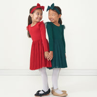 Two children posing together wearing matching Holiday Red and Emerald flutter tutu dresses paired with coordinating luxe bow headbands