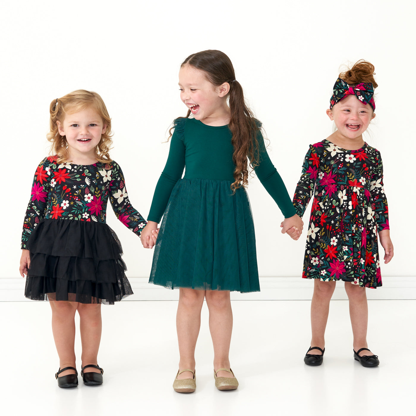Three children posing together wearing holiday play capsule dresses. One child is wearing an Emerald Flutter tutu dress. Another child is wearing a Berry Merry Flutter Tutu Dress with bloomer. The last child is wearing a Merry Berry twirl dress with bodysuit paired with a matching Berry Merry luxe bow headband
