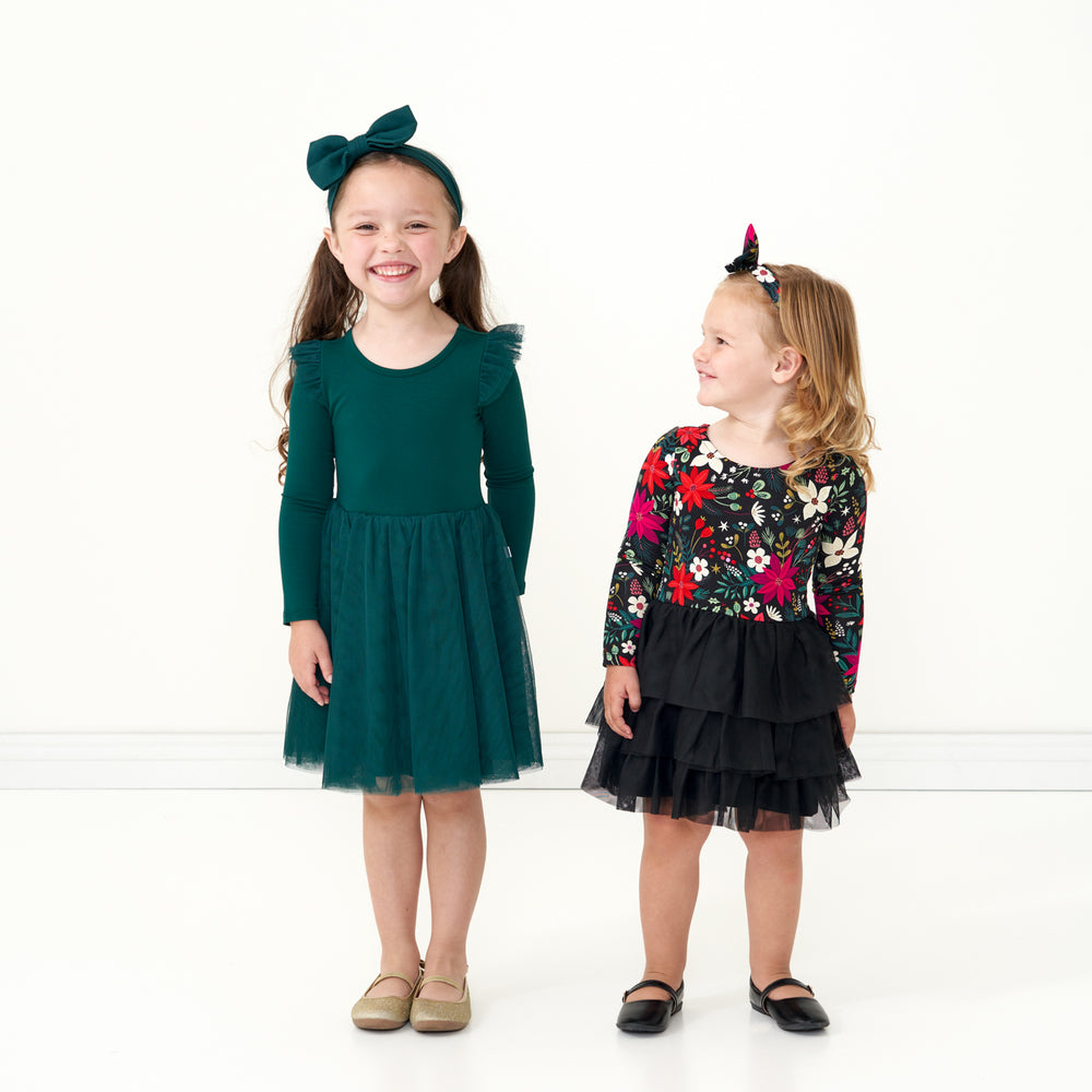 Two children posing together wearing holiday play capsule dresses. One child is wearing an Emerald Flutter tutu dress paired with a matching emerald luxe bow headband. The other child is wearing a Berry Merry Flutter Tutu Dress with bloomer paired with a matching Berry Merry luxe bow headband