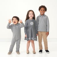 Three children posing together wearing coordinating holiday play styles. One child is wearing a Heather Charcoal Stripes polo romper, one is wearing a Snowflake sweater dress, and the last child is coordinating wearing a Heather Charcoal Stripes polo shirt.