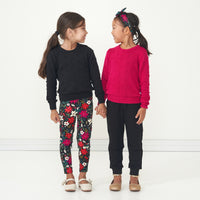 Two children holding hands together wearing holiday play capsule outfits. One child is wearing Berry Merry leggings paired with a Black Pom Pom sweater. The other child is wearing a Mixed Berry Pom Pom Sweater paired with Black Joggers and a Berry Merry luxe bow headband 