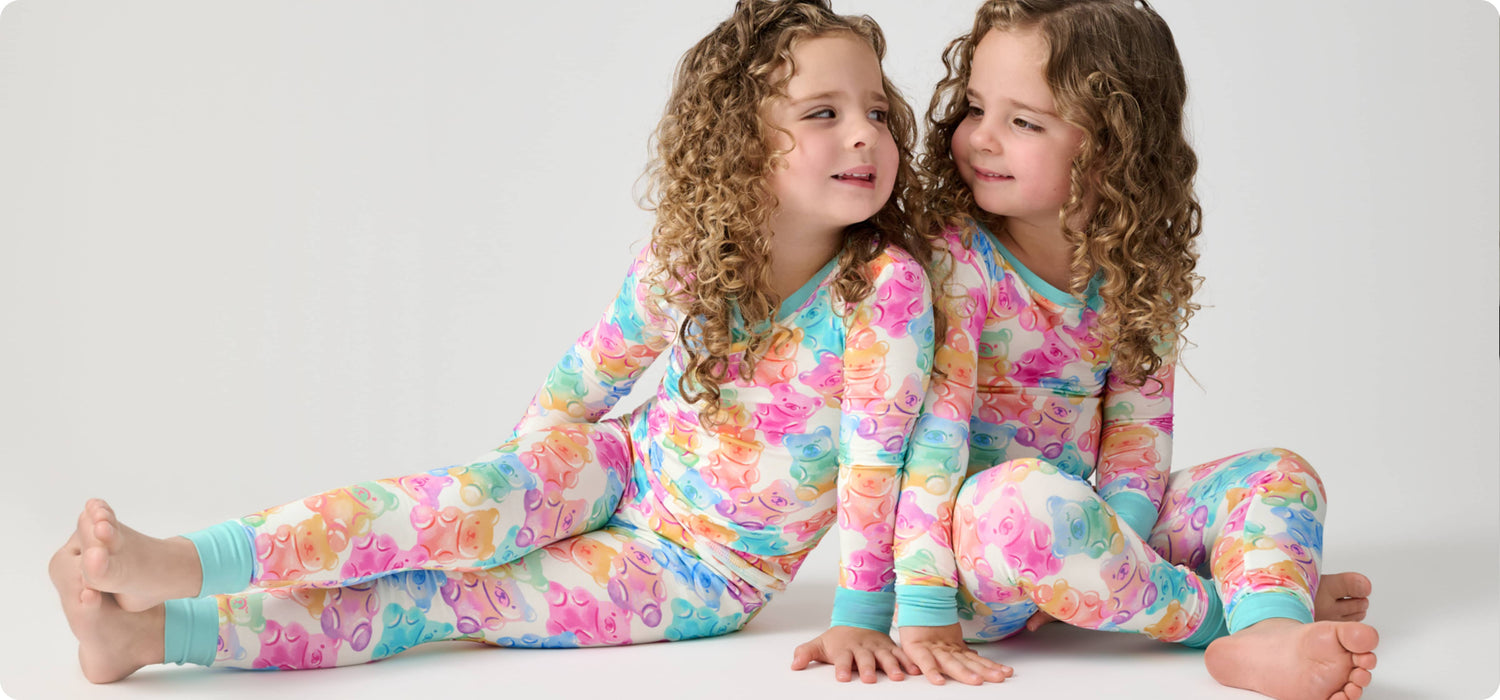 Two young girls with curly brown hair sitting side-by-side on a light gray studio floor, wearing matching long-sleeve pajamas with a colorful pastel butterfly pattern.
