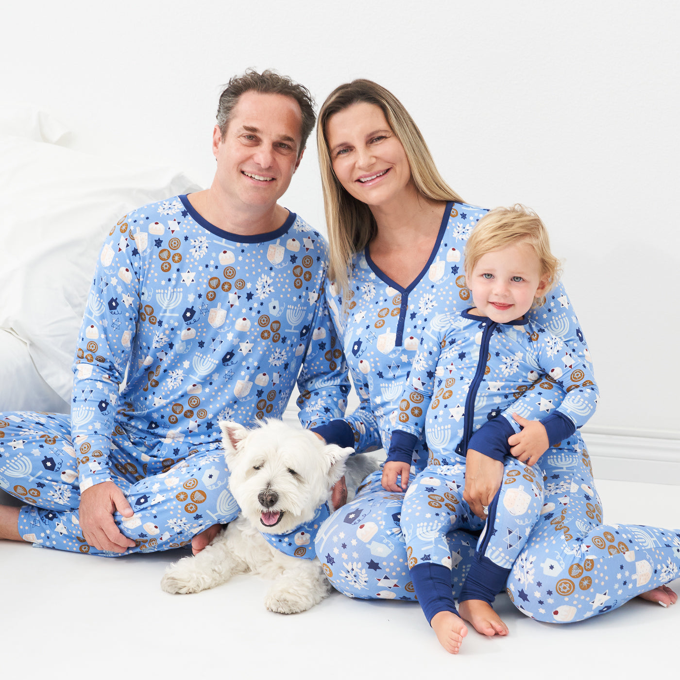 Family of three posing together wearing Hanukkah Lights and Love pajama sets. Dad is wearing a Hanukkah Lights and Love men's pajama top and matching men's pajama bottoms. Mom is wearing Hanukkah Lights and Love women's pajama top and matching pajama bottoms. Child is wearing a Hanukkah Lights and Love zippy and their dog is wearing a Hanukkah Lights and Love pet bandana