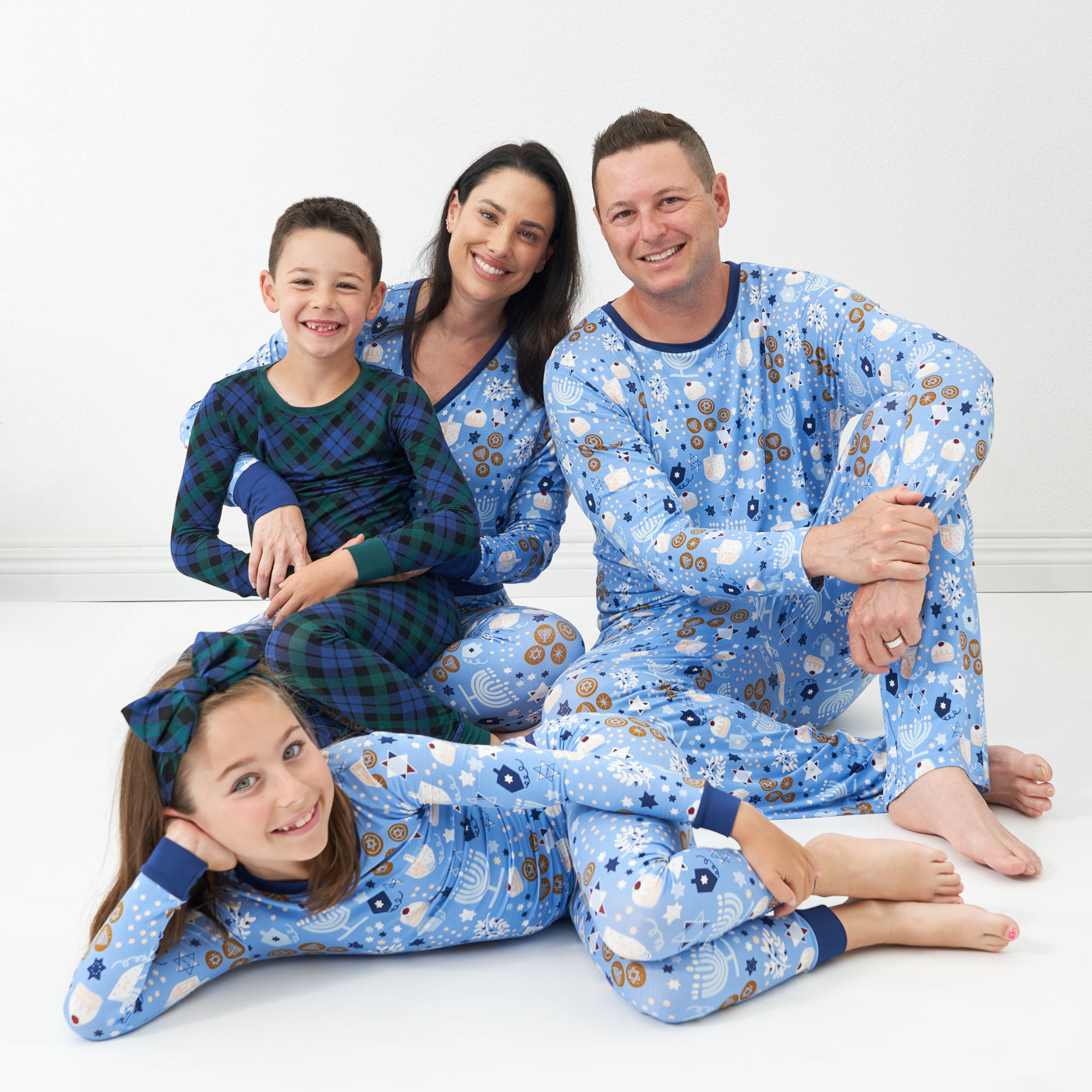 Family of four posing together. Mom is wearing Hanukkah Lights and Love women's pajama top and matching women's pajama bottoms. Dad is wearing Hanukkah Lights and Love men's pajama top and matching men's pajama bottoms. Their daughter is wearing a Hanukkah Lights and Love two piece pajama set paired with an Emerald Plaid luxe bow headband. Their son is coordinating wearing Emerald Plaid two piece pajama set